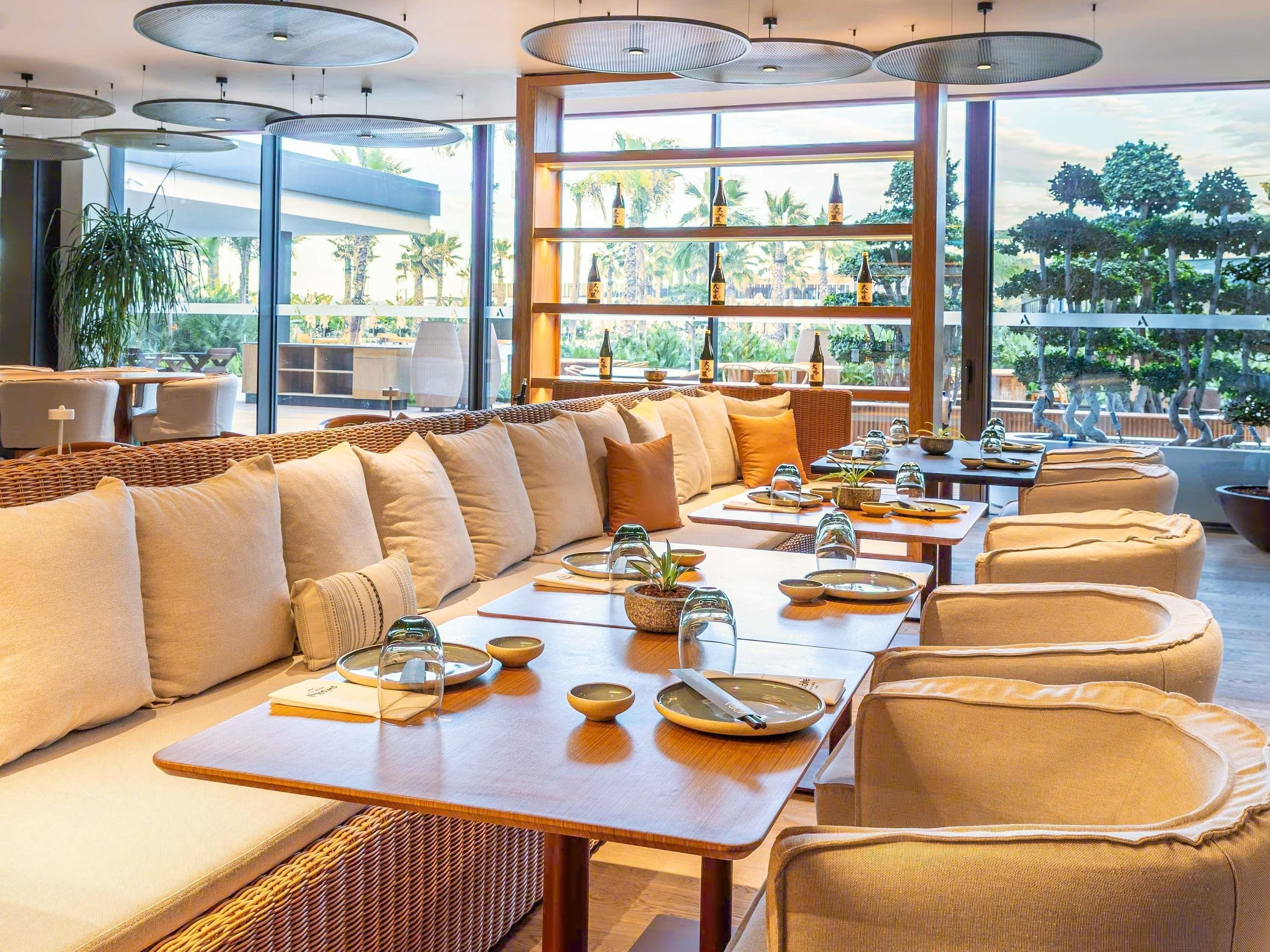 Un restaurant doté d’une longue table à manger, de sièges confortables et de grandes fenêtres donnant sur des palmiers.