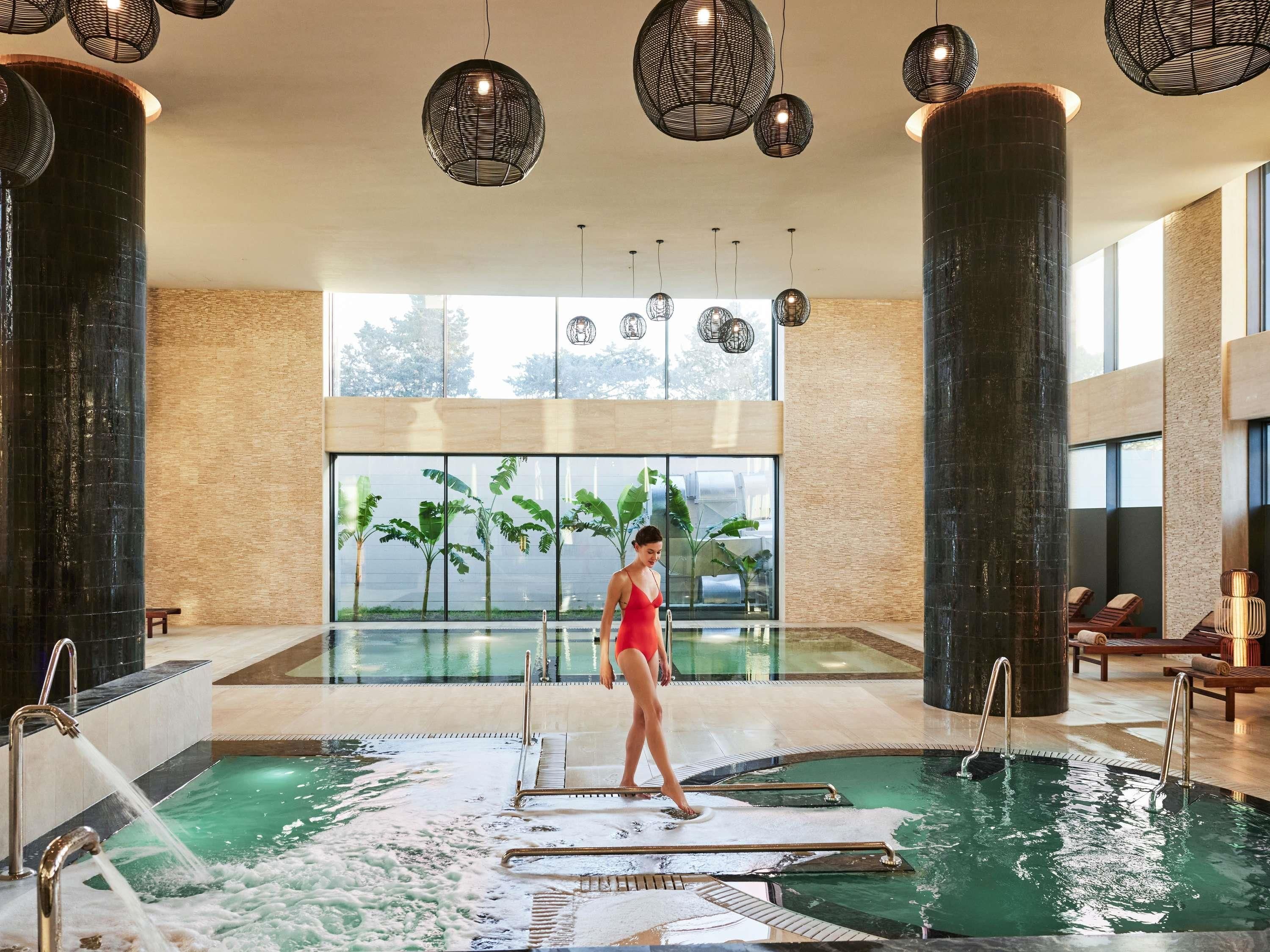 Une grande piscine intérieure où l’on aperçoit une personne portant un maillot de bain rouge, entourée de grandes colonnes, d’éclairages suspendus modernes et offrant une vue sur la verdure à l’extérieur.
