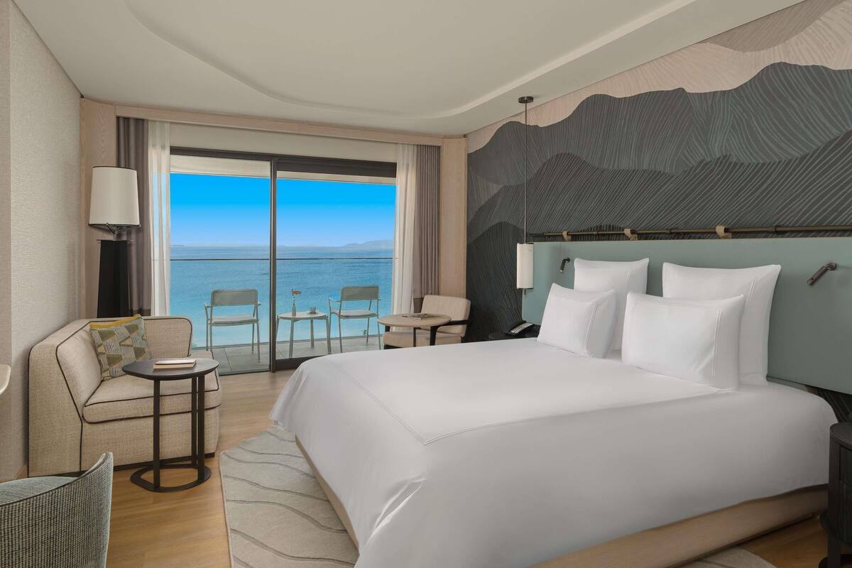 A hotel room with a bed, a sofa, a small table, a chair, and a view of the sea.