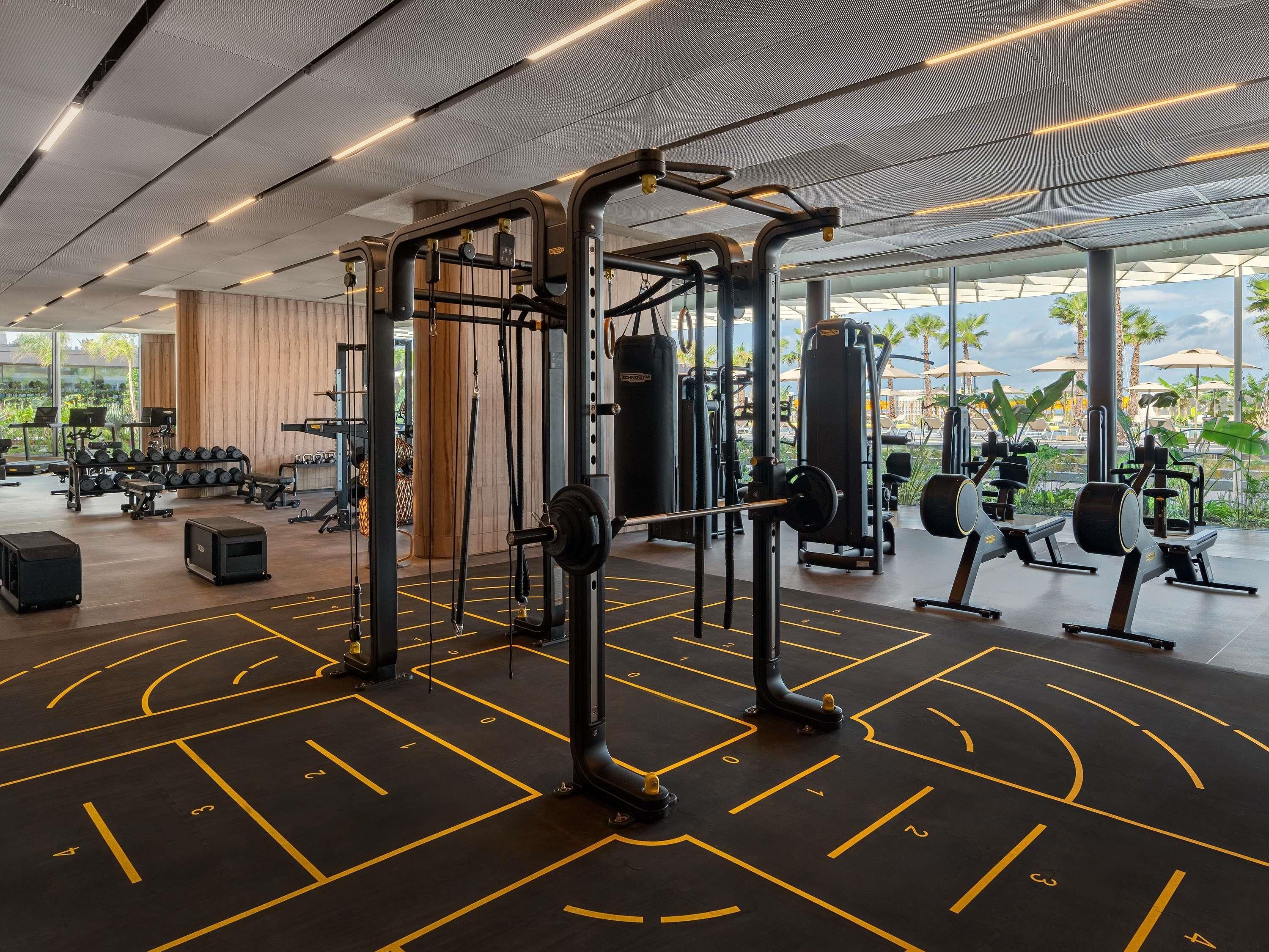 Ein moderner Fitnessraum mit Krafttrainingsgeräten, einem großen Spiegel und Blick auf Palmen durch große Fenster.