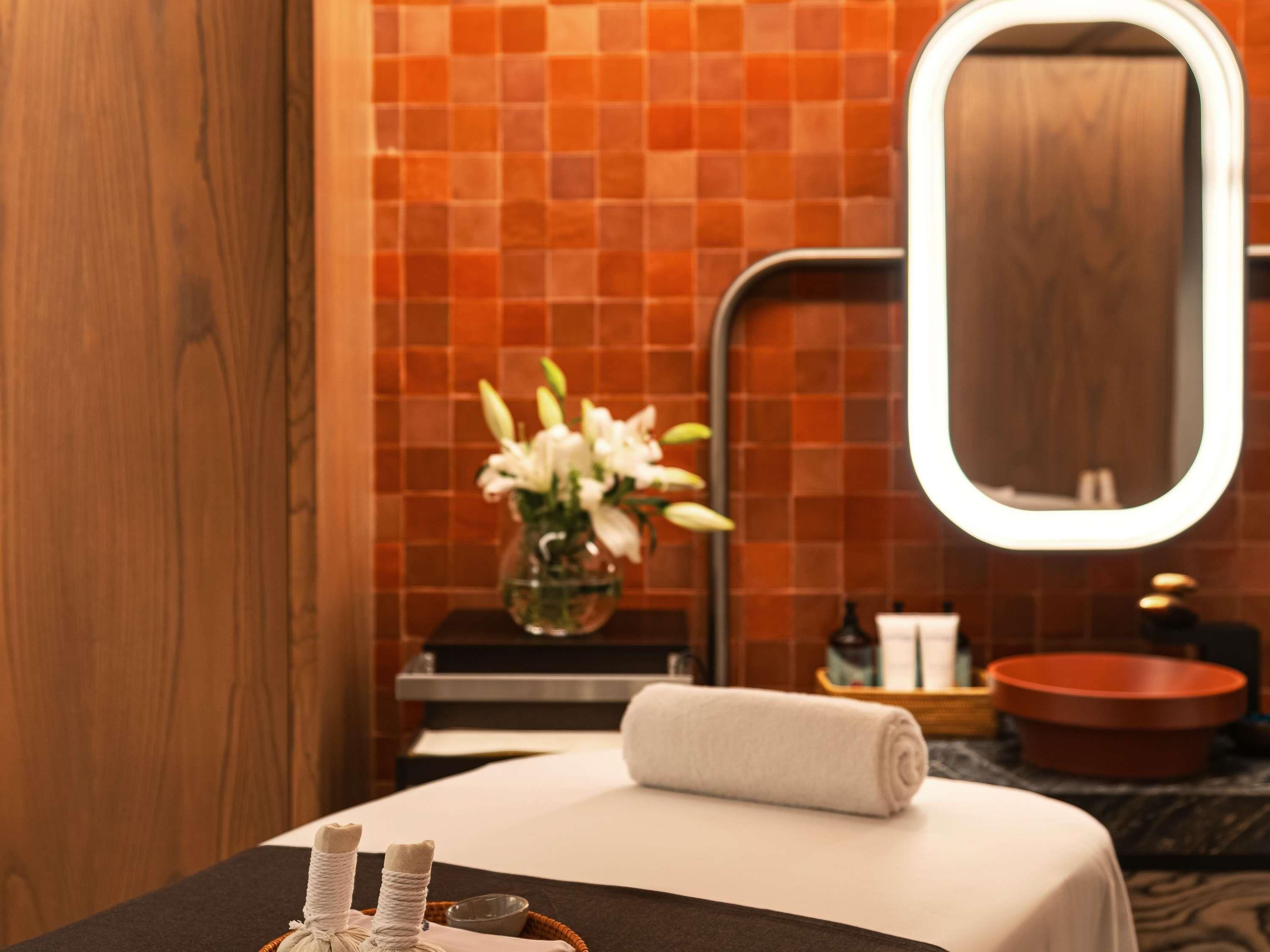 A bathroom with a tiled wall, a mirror, a sink, a towel, and a vase with flowers.