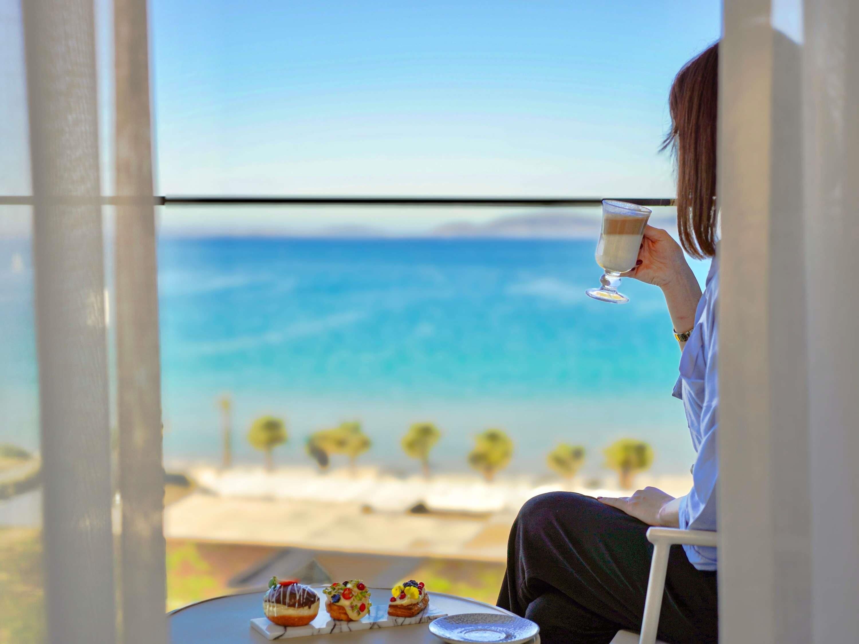 A person seated at a table with a glass of drink and three pastries, overlooking a beach with the sea and palm trees.