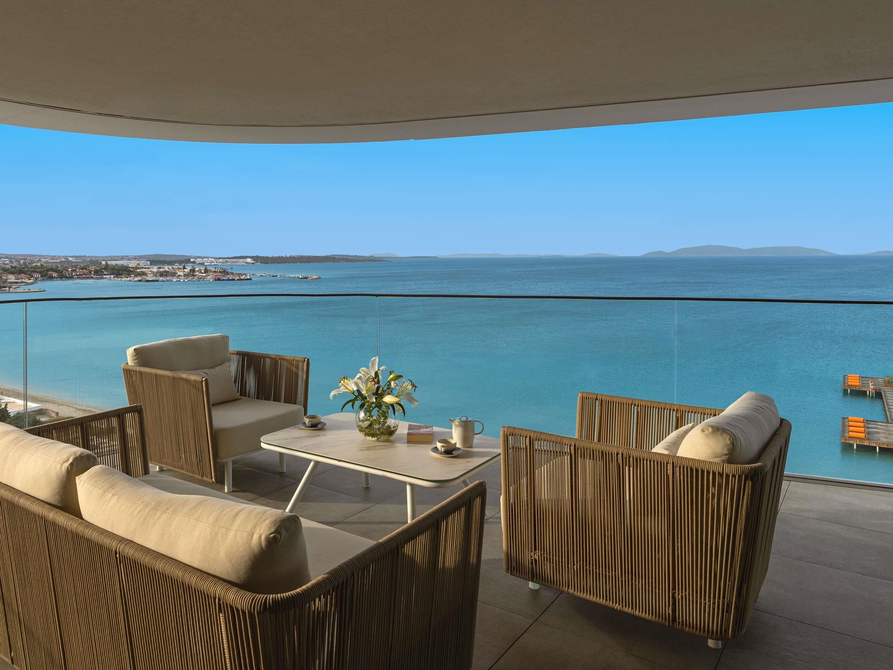 A balcony with wicker furniture, a vase with flowers, and a clear view of the sea and distant land.