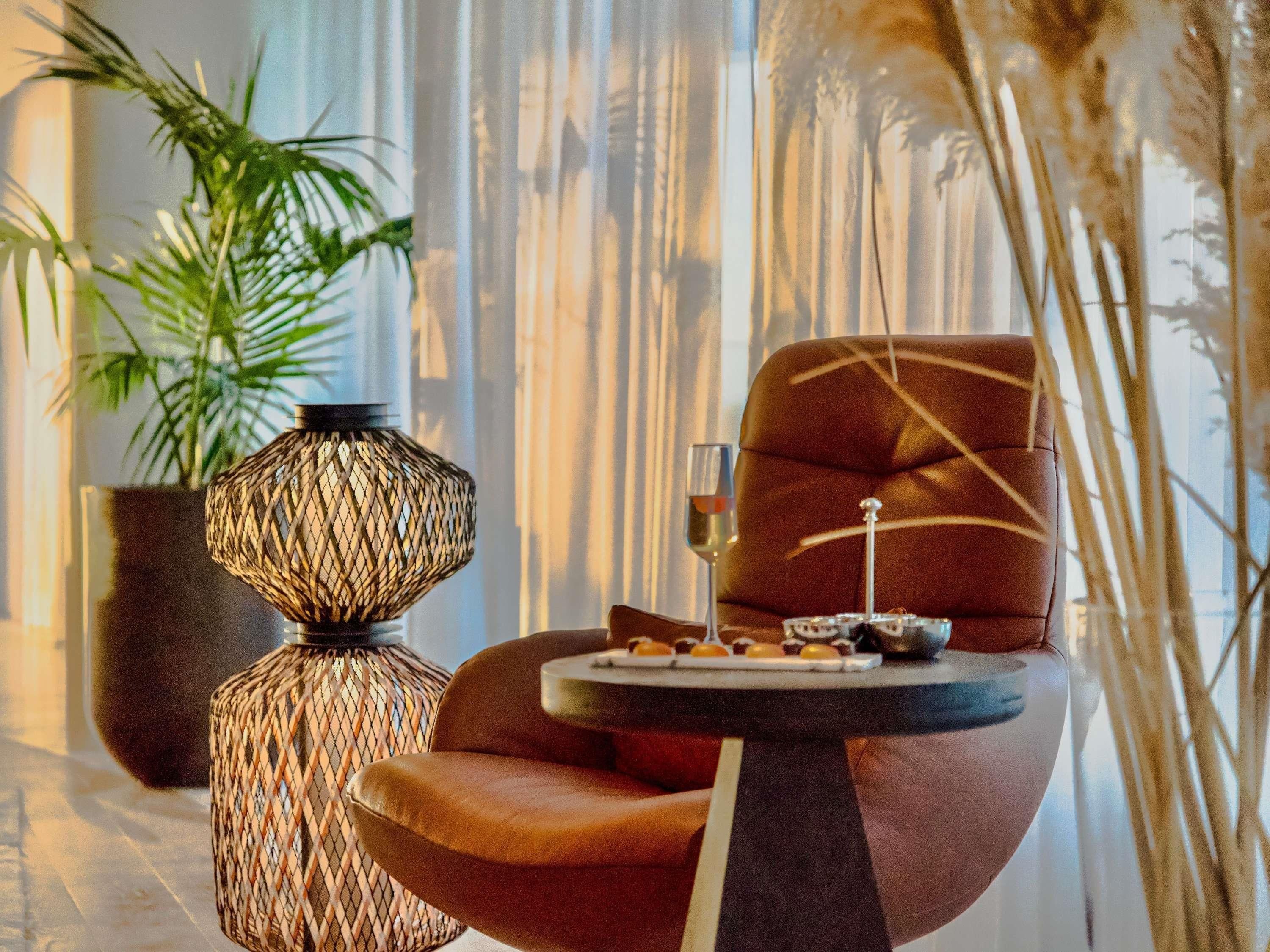 Un interior moderno con un sillón de cuero, una lámpara trenzada, una copa de vino y una pequeña mesa con bombones.