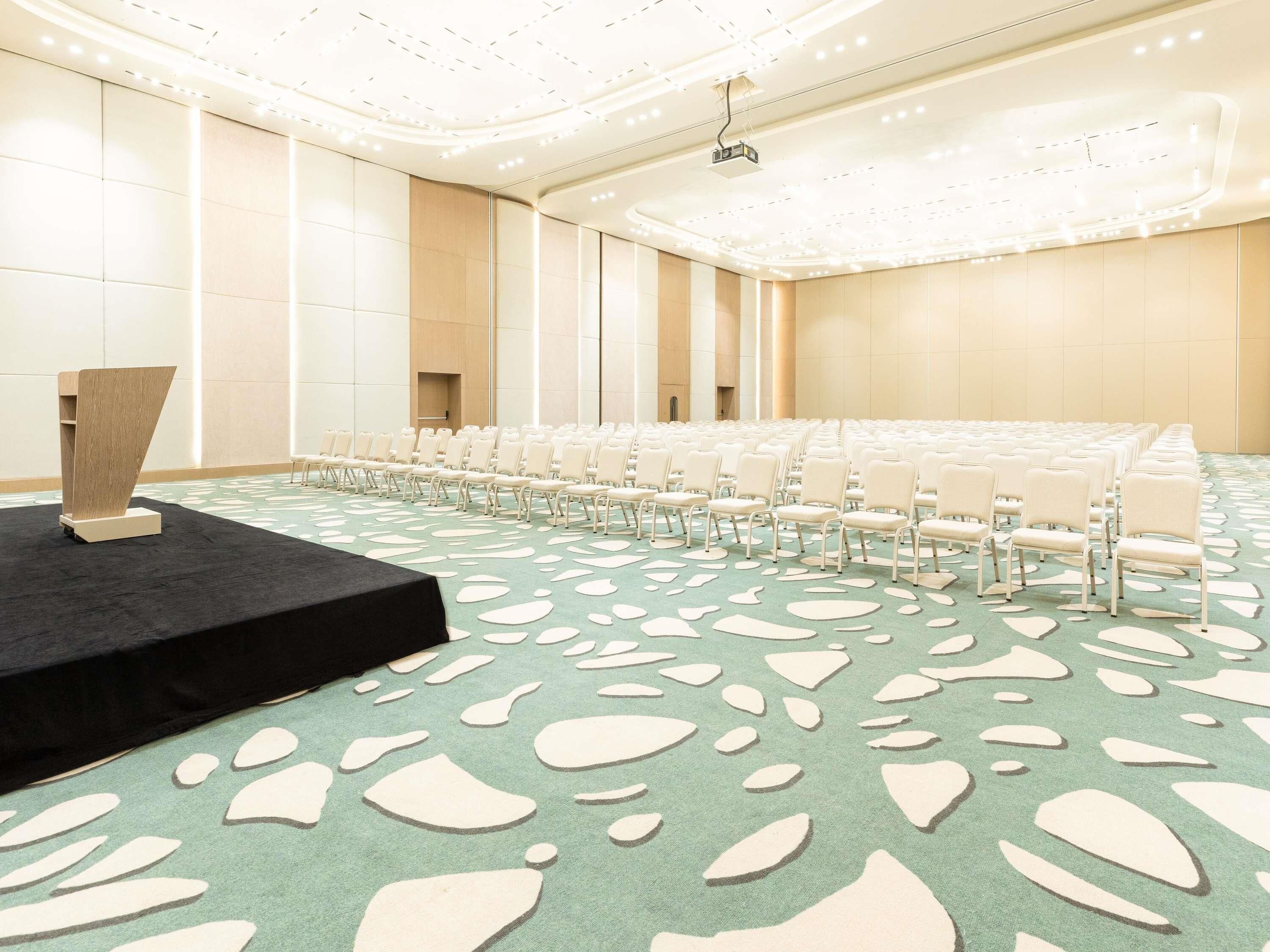 A conference room with rows of white chairs, a stage with a podium, and a patterned carpet.