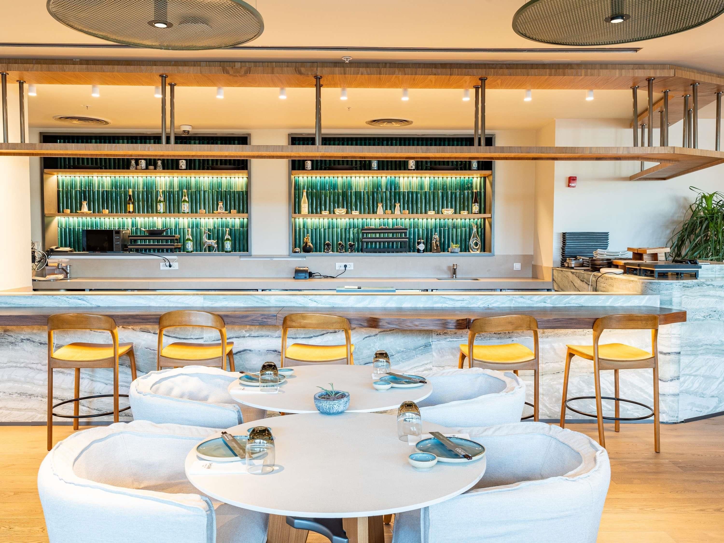 Zona de bar moderna con fondo de bambú verde, encimeras de mármol y mesas redondas con sillas.