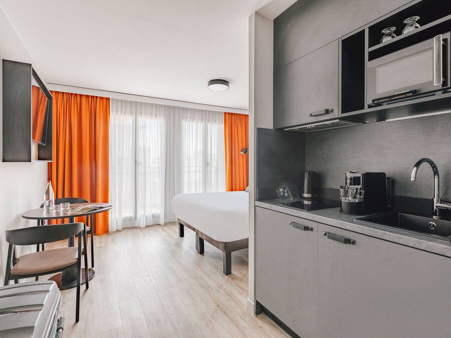 Une chambre d’hôtel moderne comprenant un lit, une petite table à manger avec des chaises, un coin cuisine équipé d’un micro-ondes et une grande fenêtre avec des rideaux orange.