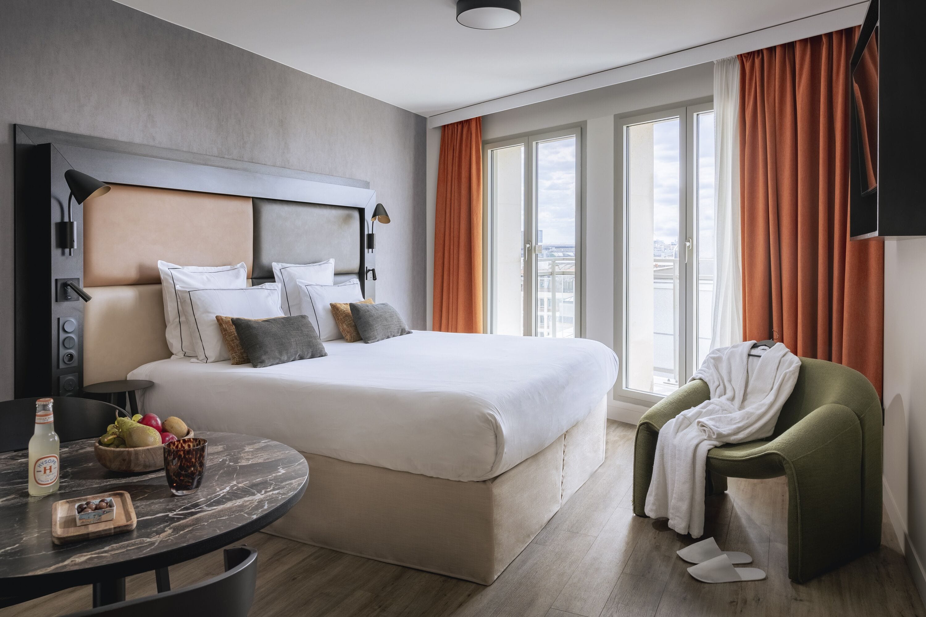 A modern hotel room with a large bed, a round table with breakfast items, a green armchair, and a city view through the windows.