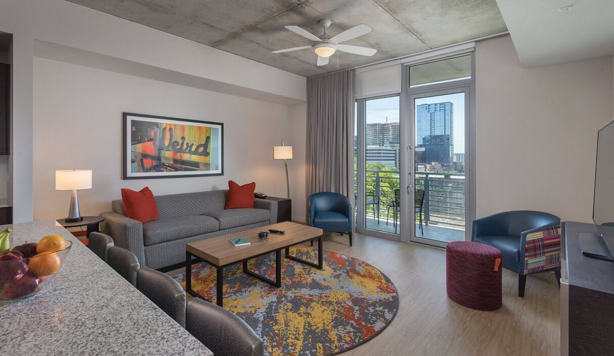 Una sala de estar moderna con un sofá gris, almohadas rojas, una mesa de centro de madera y un tapete colorido. Se observa un sillón azul, un reposapiés rojo y un escritorio con una lámpara. Se ve un ventilador de techo, y la habitación tiene ventanas amplias con vistas a la ciudad.