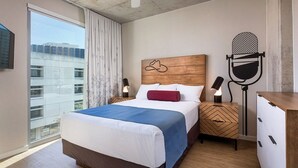 A hotel room with a large bed, a wooden headboard, a nightstand with a microphone, and a view of a building through a window. - 2BD/Austin - Full Kitchen, W/D, Balcony, Fire Pit! (Austin)