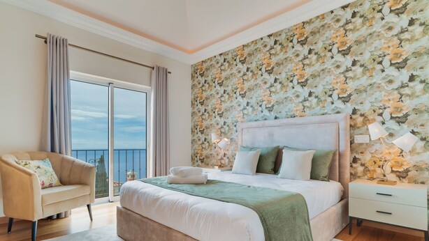 A bedroom with a large bed, a nightstand, a chair, and a view of the sea through a balcony. - Villa Gasparini (Funchal)