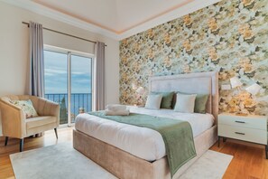 Un dormitorio con una cama grande, una mesita de noche, una silla y vistas al mar desde un balcón.