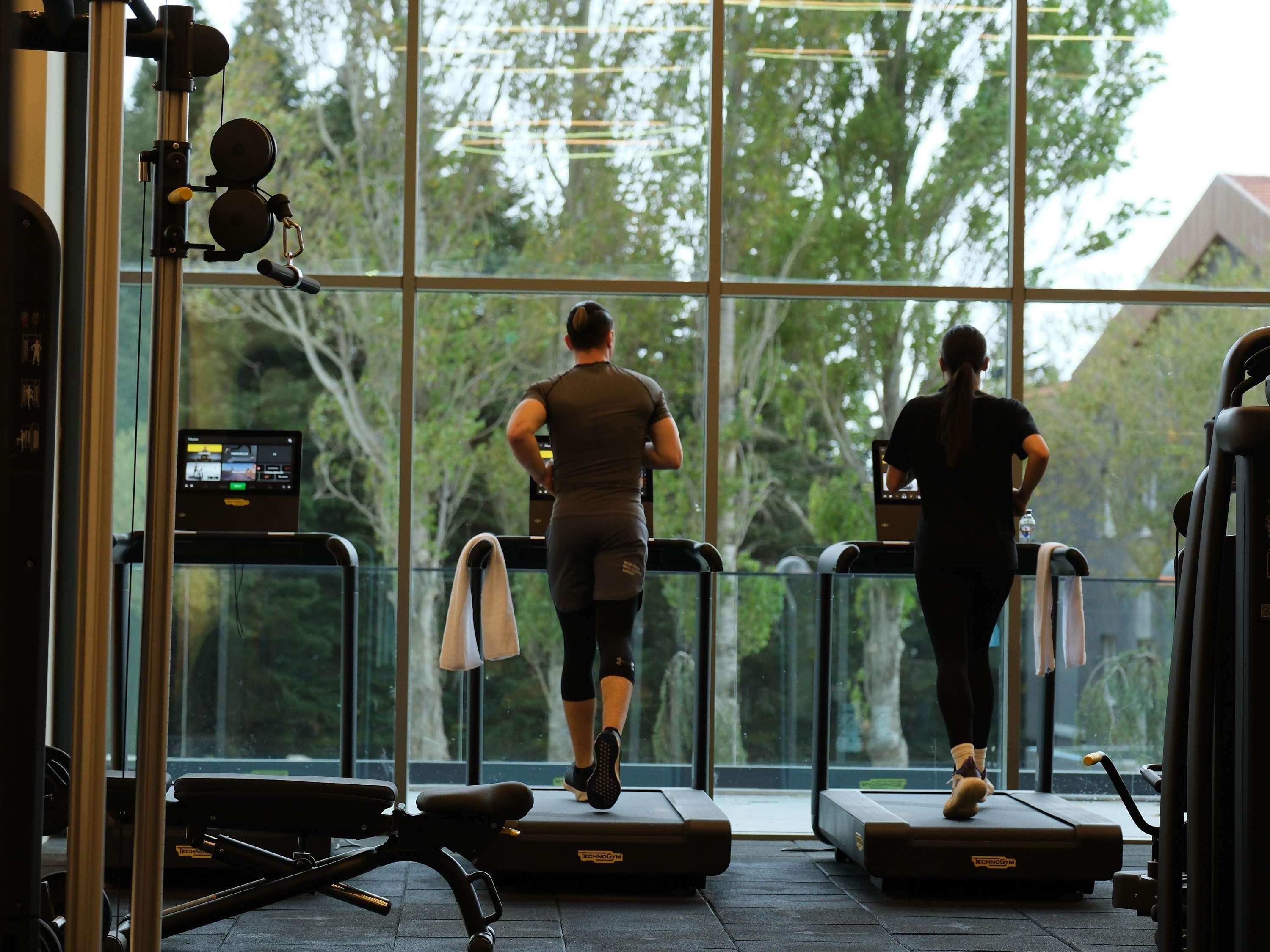 Un gimnasio con cintas de correr, equipo de ejercicio y amplios ventanales que ofrecen vistas a árboles y edificios.