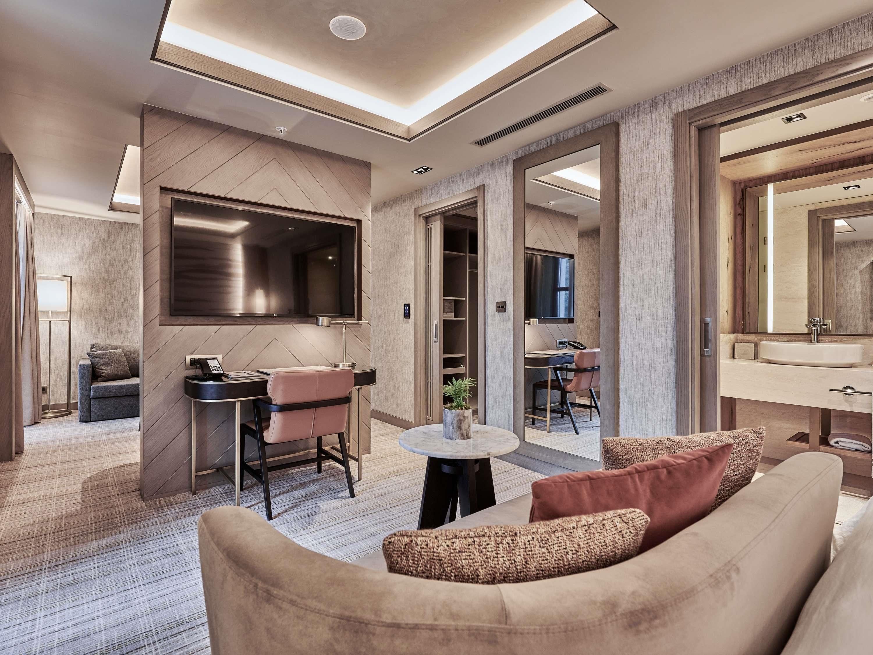 A modern hotel room with a sofa, a desk with a chair, a TV, a large mirror, and a walk-in closet.
