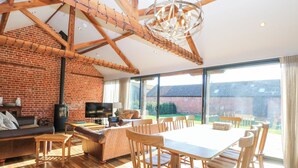 A spacious living room with a large wooden table, comfortable seating, a fireplace, and a large window offering a view of the outdoors. - Shorthorn Barn (Norwich)