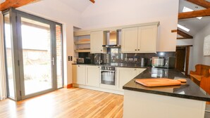 A modern kitchen with wooden beams, a large island, and a sliding glass door leading to a patio. - Shorthorn Barn (Norwich)
