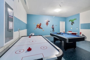 A recreational room with an air hockey table, a pool table, and a blue wall with superhero-themed decorations.