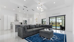 A modern living room with a grey sectional sofa, a coffee table, and a blue rug with a compass design. There is a kitchen area with white cabinets and a large island. A glass door leads to a pool area.