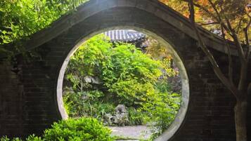 Un arco de piedra con un vano circular, que da acceso a un jardín con diversas plantas y una estructura de techo tradicional con tejas al fondo.
