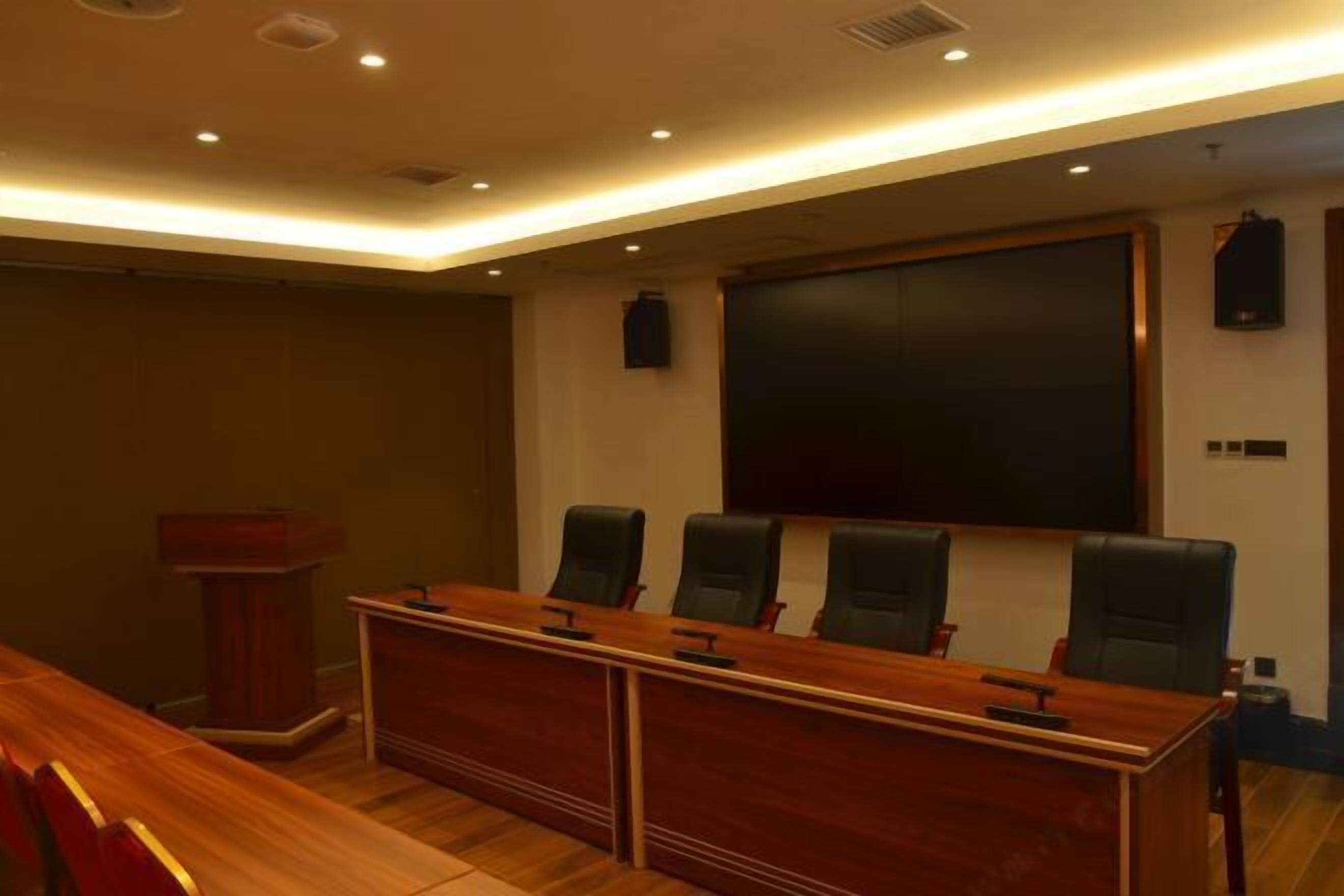 Un salón de conferencias con una larga mesa de madera, cuatro sillas negras, un atril y una pantalla grande en la pared.