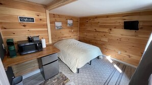 A compact room with wooden walls, a bed, a kitchenette with a microwave, and a coffee bar sign.