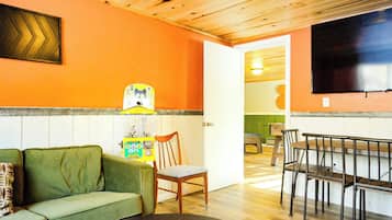 Un salon chaleureux avec un canapé vert, une table à manger en bois et des chaises. Un téléviseur est fixé au mur, et une porte donne accès à une autre pièce.