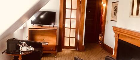 A hotel room with a large bed, a television on a wooden stand, a small table with a chair, and a fireplace.
