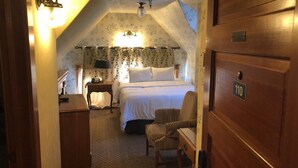 A cozy bedroom with a bed, a chair, a nightstand, and a lamp. The room has a wooden door with a number plate.