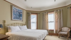A bedroom with a bed, a chair, a painting, and windows with curtains. - Blocks from Ocean - KING BEDROOM SUITE (PACIFIC GROVE)