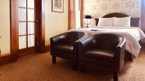 A hotel room with a large bed, two leather armchairs, a nightstand with a lamp, and a floral wallpaper pattern.