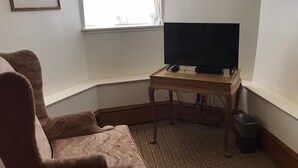 Una habitación de esquina con un ventanal grande, un televisor sobre un soporte de madera, una alfombra estampada y un cuadro en la pared.