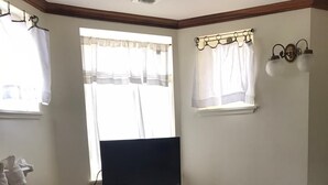 Una habitación de hotel con una cama, un mueble de televisión de madera con un control remoto, un cubo de basura pequeño y una ventana con cortinas.