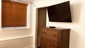 A room with a large window with blinds, a wooden dresser with drawers, and a flat-screen TV mounted on the wall. - Queen Bed with Bunk bed (PACIFIC GROVE)