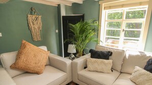 Een woonkamer met een beigekleurige zithoek, een macramé wanddecoratie, een potplant en een raam met uitzicht op groen.