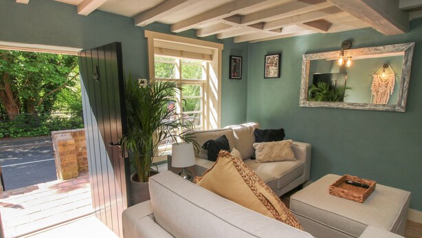 Een woonkamer met een bank, een salontafel en een bloempot. Er is een raam met zonwering en een deur naar buiten.