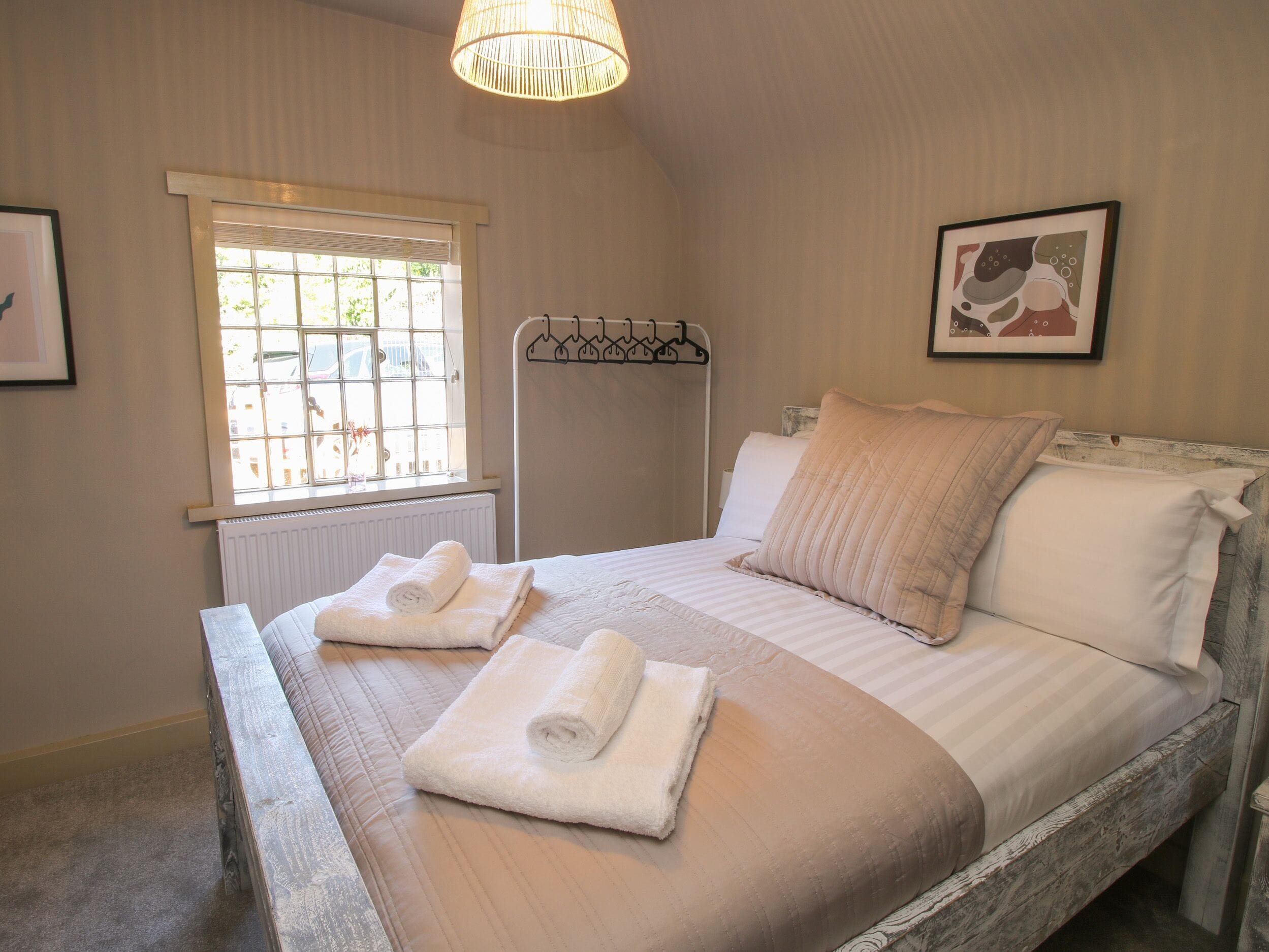 A neatly made bed with rolled towels, a radiator, a window with blinds, and a hanging rack.