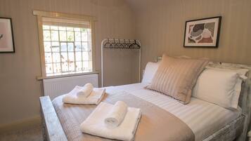 A neatly made bed with rolled towels, a radiator, a window with blinds, and a hanging rack.