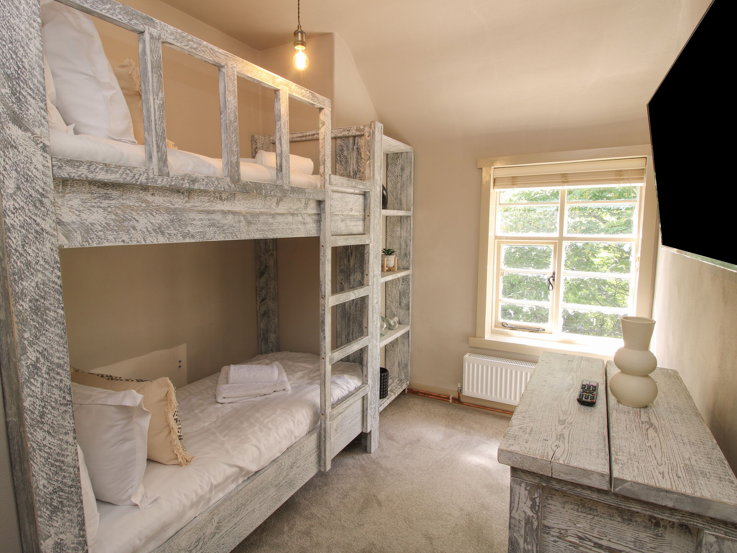 A bunk bed room with a window, a radiator, and a small table with a vase and a remote control.