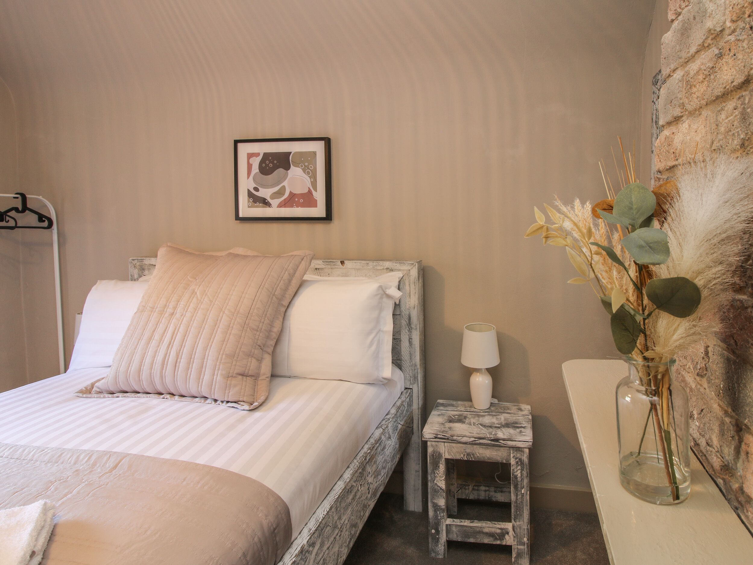 Un dormitorio con cama, una mesita de noche con lámpara, un cuadro en la pared y un florero con plantas secas.