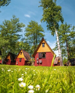 A row of red and yellow wooden houses with a grassy foreground and trees in the background. - Kemp Sluníčko (Doksy)