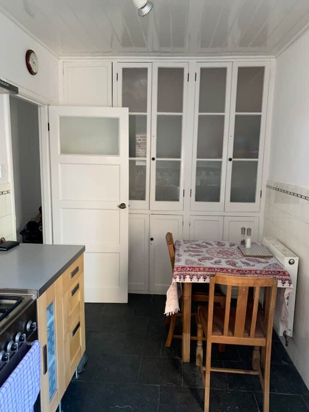 Uma cozinha com armários brancos, uma mesa de madeira com toalha de mesa estampada e um relógio na parede.