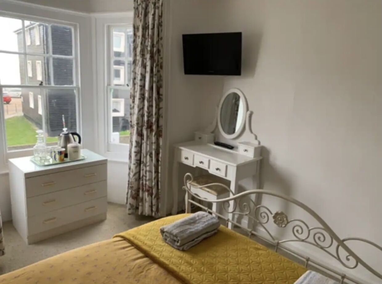 A bedroom with a bed, a dresser, a vanity with a mirror, and a TV mounted on the wall.
