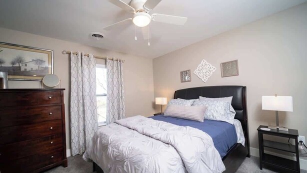 Un dormitorio con cama, cómoda, mesita de noche, ventilador de techo y ventana con cortinas.