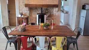 Un coin repas rustique avec une table en bois, quatre chaises, un mur en briques, un poêle à bois et, au fond, un espace cuisine.