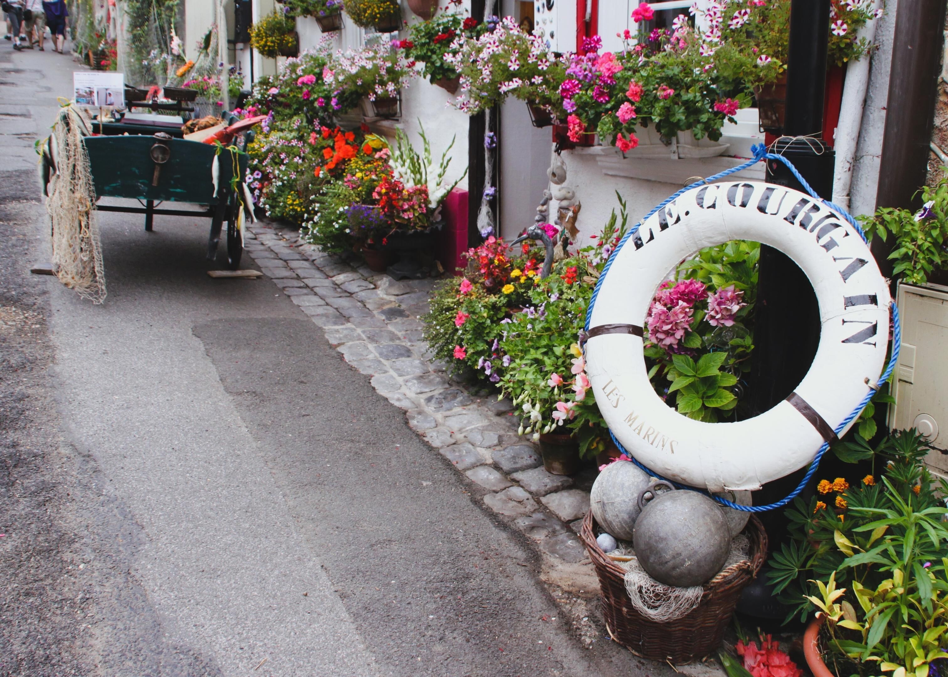 En gata med en rad blomlådor och en livboj med texten ”LES MARINS” på trottoaren.