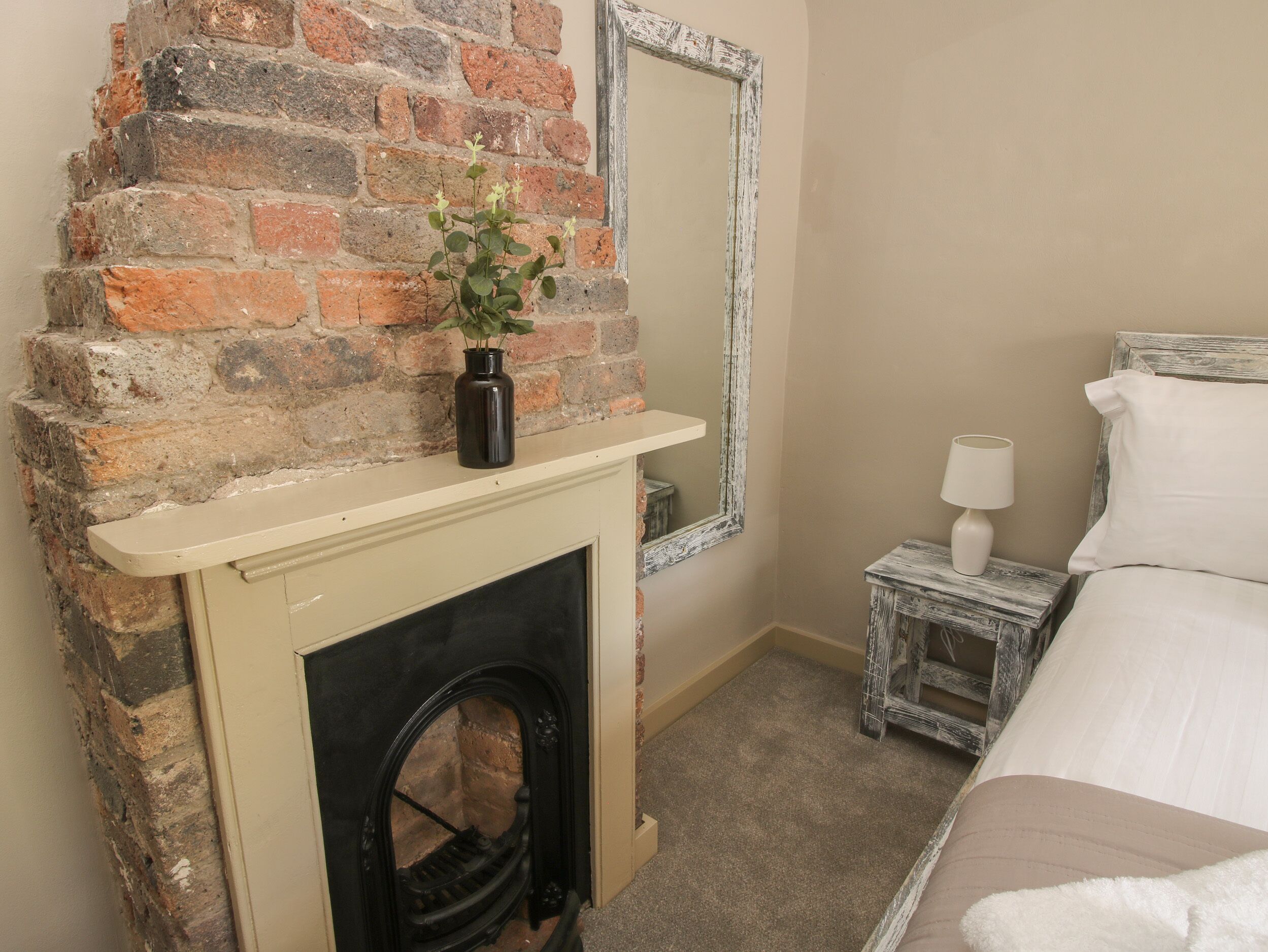 A bedroom with a brick fireplace, a small bedside table with a lamp, and a bed with white bedding.