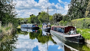 A narrow canal with three boats docked along the banks, surrounded by lush greenery and a clear blue sky. - Boutique Boat Stays Contemporary Barge (Bath)