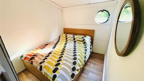 A small, single-bed room with a wooden headboard, a round mirror on the wall, and a window with a view of greenery. - Boutique Boat Stays Contemporary Barge (Bath)