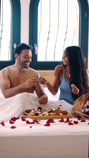 A couple sitting on a bed with a tray of food and rose petals, in a room with large windows and a dark-colored wall.