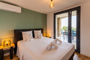 Ein Hotelzimmer mit einem großen Bett, zwei Nachttischen mit Lampen und Blick durch eine Glastür auf einen Außenbereich mit Tisch und Stühlen.